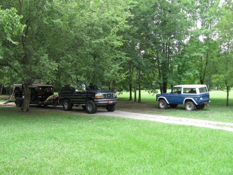 1967 Ford Mustang - A blue Ford Bronco sits in a grassy area near trees, while a black Ford F-Series truck tows a trailer with a Jeep.