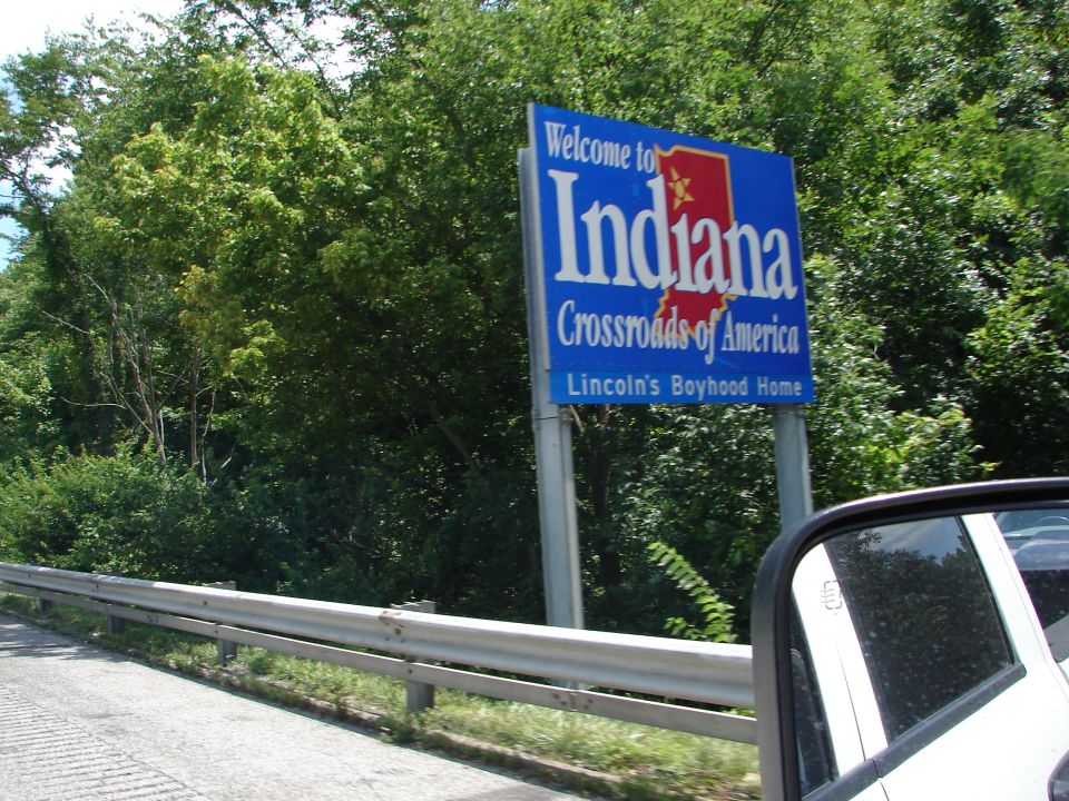 Welcome to Indiana sign on roadside with trees and guardrail. Part of a vehicle is visible in the foreground