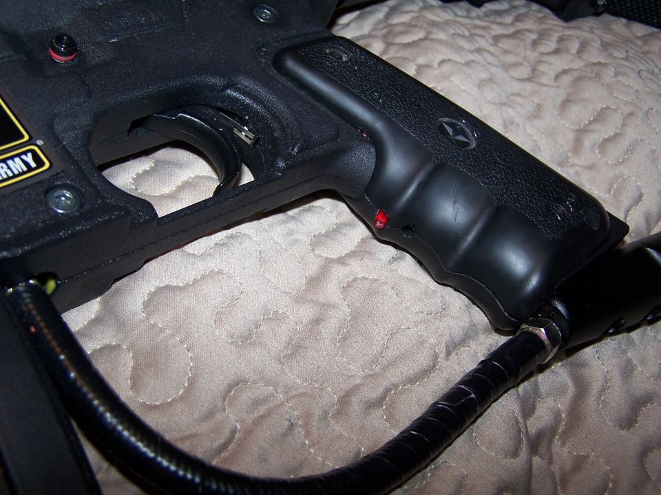 Close-up of a black paintball gun's grip and trigger assembly. A black hose connects to the bottom