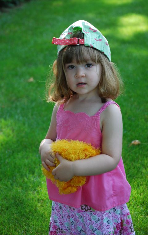 2000 Ford Crown Victoria - A young child stands in a grassy yard, holding a fluffy yellow toy and wearing a cap with a cherry pattern.