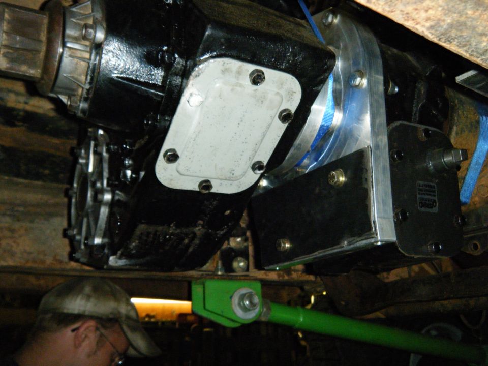 Close-up of black and silver drivetrain parts, including a transfer case, with a green suspension component and a person's head in the lower left. Text 'SUPERMOTORS' visible