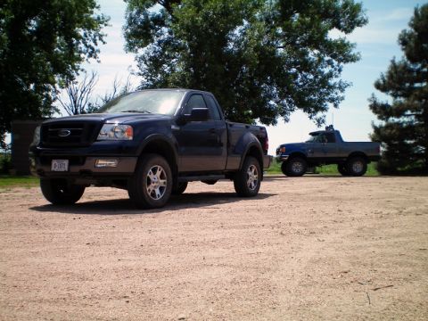 2005 Ford F-150 - Two pickup trucks are parked on a gravel lot with trees in the background.