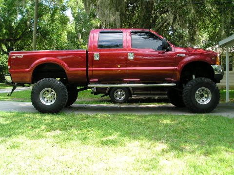1978 Ford Bronco - A lifted red pickup truck is parked on a paved surface with grass and trees in the background.