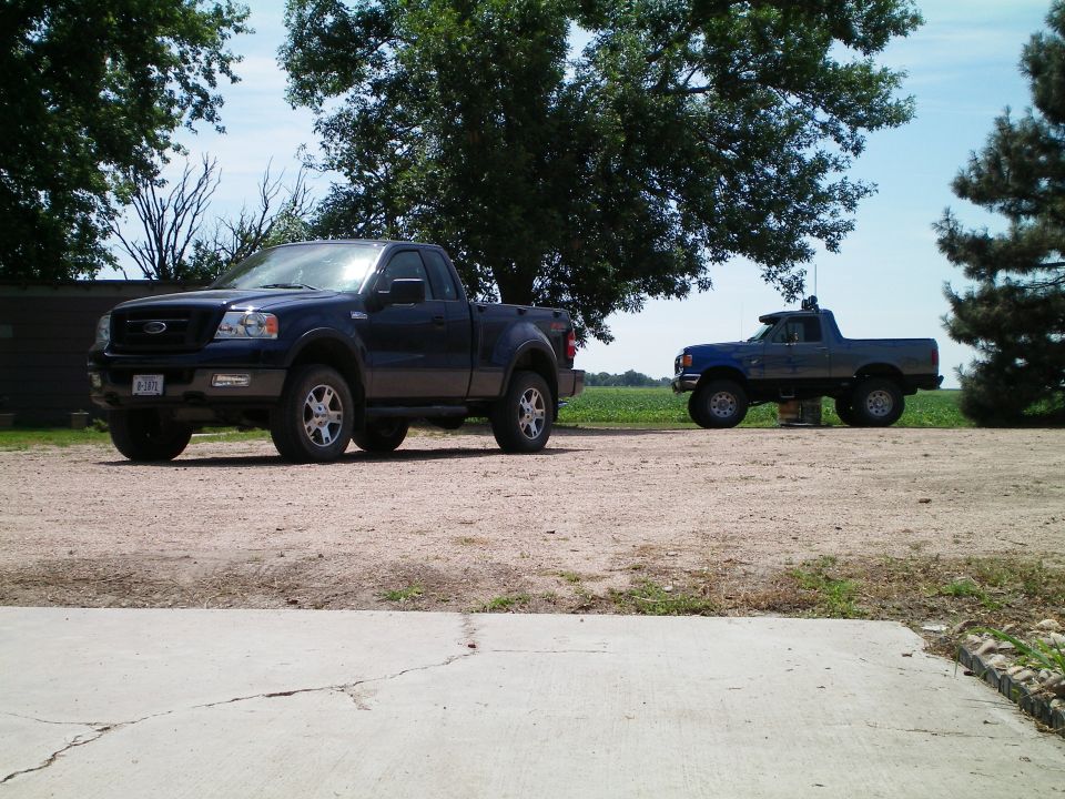 Dark blue Ford F-150 regular cab parked on gravel with an older lifted blue Ford truck behind it on a farm.