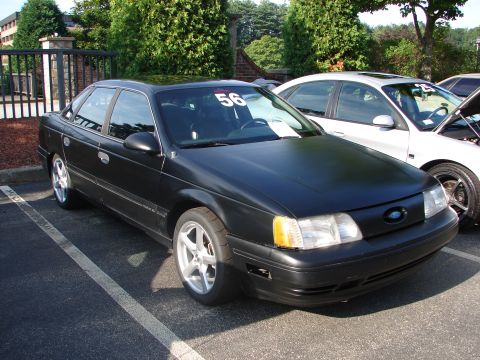 1992 Ford Taurus - A black 1992 Ford Taurus is parked in a lot, displaying racing numbers on its windshield.