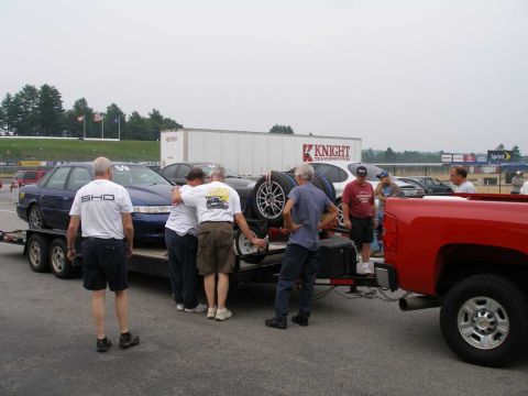 1997 Ford Taurus - A group of people gather around a blue Ford Taurus SHO being loaded onto a trailer, with a red pickup truck in the foreground.