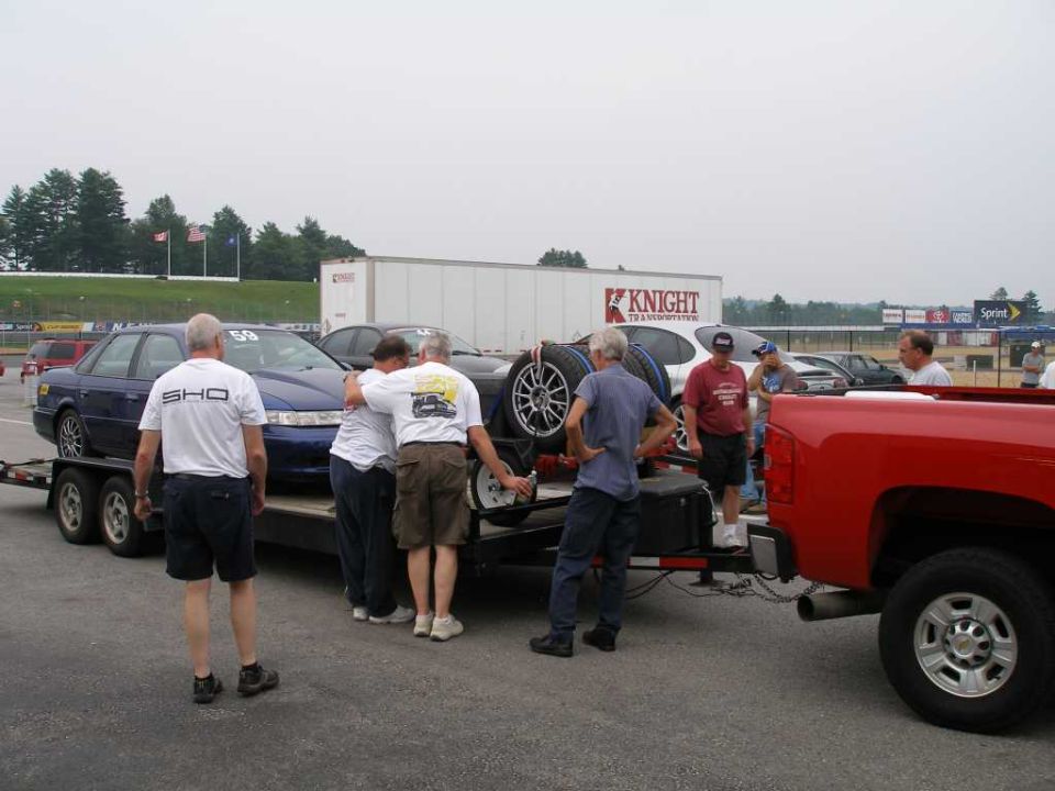 Blue Ford Taurus SHO on trailer, people around it, red pickup truck nearby