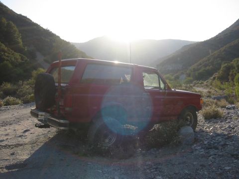 1988 Ford Bronco - Red full-size Ford Bronco parked side-on across a rocky dirt trail in a canyon, backlit by the sun cresting the ridge and throwing a lens f…