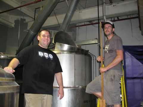1985 Ford LTD Crown Victoria - Two men pose for a photo in a brewery, with large industrial brewing equipment behind them.