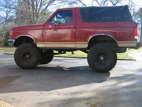 1990 Ford Bronco - Side view of a lifted 1990 Ford Bronco with a red body and tan trim, equipped with large off-road tires.