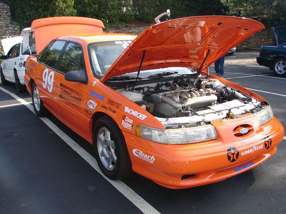 Orange 1992 Ford Taurus race car with hood open, showing engine. Number 99 on door. Other cars visible in background