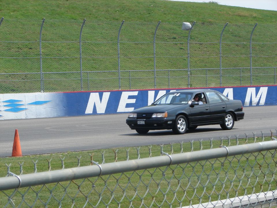 Black 1992 Ford Taurus sedan driving on a race track with a driver visible inside