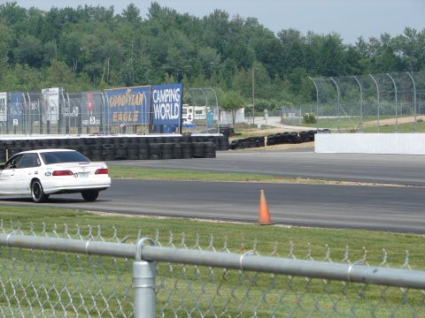 1992 Ford Taurus - A white 1992 Ford Taurus is seen on a race track, with a backdrop of trees and advertising banners.