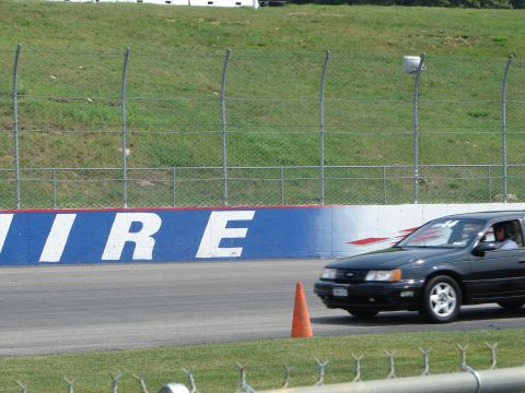 1992 Ford Taurus - A black 1992 Ford Taurus navigates a race track, passing a traffic cone near a branded wall.