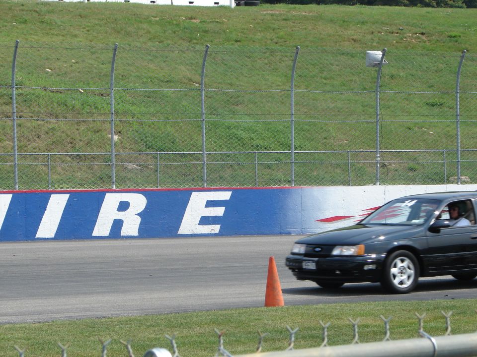 Black 1992 Ford Taurus sedan on a race track, front 3/4 view, driver visible