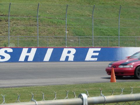 1992 Ford Taurus - A red car with a black hood stripe drives on a race track next to a wall with 'SHIRE' painted on it.