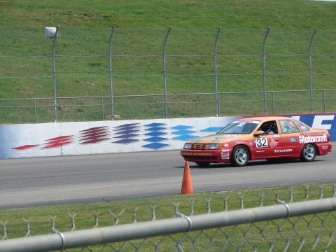 1992 Ford Taurus - Racing sedan with sponsor decals navigates a turn on a paved track with grassy infield and fence.