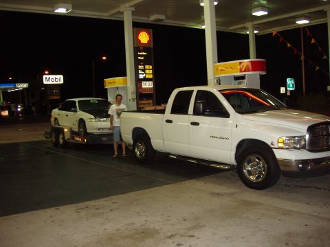 1992 Ford Taurus - A white sedan is transported on a trailer at a gas station, with a white pickup truck towing it.