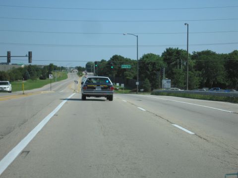 1991 Ford Crown Victoria - A highway patrol car is seen driving on a road with trees and traffic lights in the background.