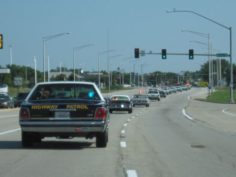 1991 Ford Crown Victoria - A line of vehicles, led by a Highway Patrol car, proceeds down a multi-lane road under a clear sky.