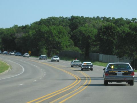 1991 Ford Crown Victoria - A line of police cars, including a 1991 Ford Crown Victoria, travel down a highway with trees lining the road.