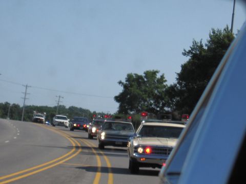 1991 Ford Crown Victoria - A motorcade of police vehicles proceeds down a two-lane highway under a clear blue sky.