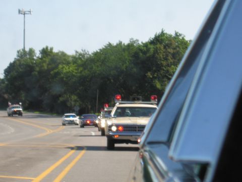 1991 Ford Crown Victoria - A line of police vehicles, some with flashing lights, travels down a highway with trees lining the sides.