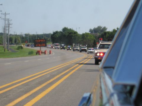 1991 Ford Crown Victoria - A convoy of vehicles, including a 1991 Ford Crown Victoria, travels down a highway with an electronic sign indicating delays.