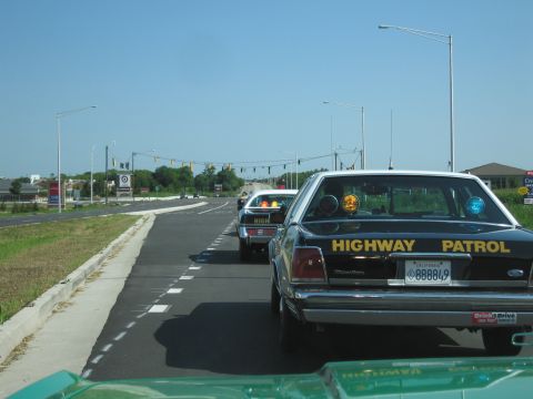 1991 Ford Crown Victoria - A line of black Highway Patrol cars, including a 1991 Ford Crown Victoria, drive down a highway on a clear day.