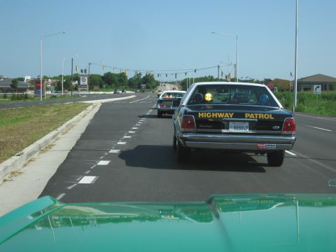 1991 Ford Crown Victoria - Two Highway Patrol cars driving on a sunny day, one directly ahead.