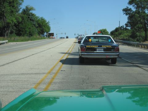 1991 Ford Crown Victoria - A black highway patrol car drives on a sunny day, with other vehicles visible ahead on the road.