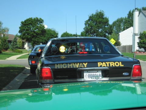 1991 Ford Crown Victoria - Two highway patrol cars are lined up on a sunny day, with houses and trees in the background.