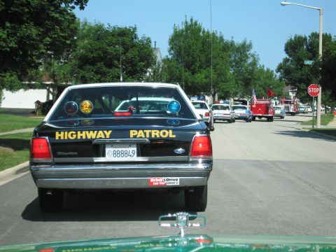 1991 Ford Crown Victoria - A line of vehicles, including police cars and fire trucks, drive down a suburban street on a sunny day.