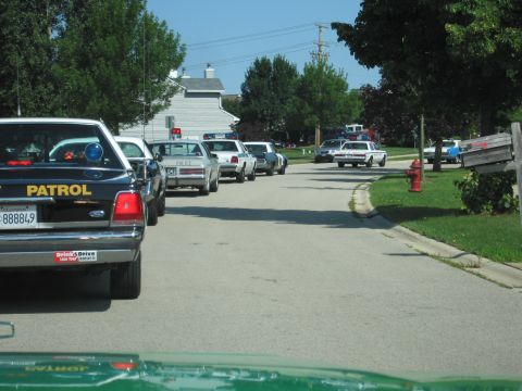 1991 Ford Crown Victoria - A convoy of police vehicles, including a black patrol car, is parked on a quiet residential street on a sunny day.