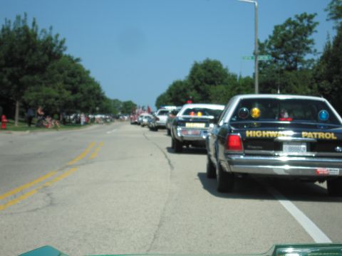 1991 Ford Crown Victoria - A line of police cars, including a 1991 Ford Crown Victoria, drive down a street lined with trees and spectators.