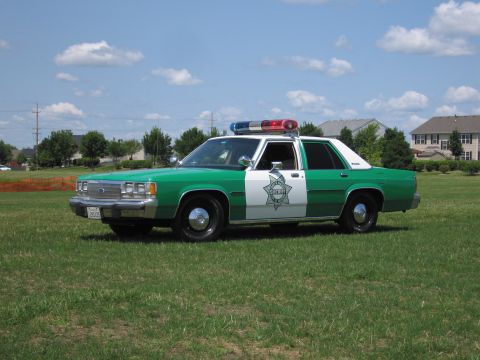 1991 Ford Crown Victoria - Sheriff's car with star emblem on door, parked on a grassy field under a cloudy sky.