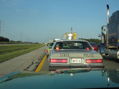 1991 Ford Crown Victoria - A silver state police car with 'STATE POLICE' on the trunk is seen in traffic behind other vehicles.