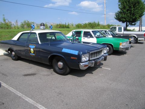 1991 Ford Crown Victoria - A line of vehicles, including a vintage police car, are parked in an outdoor lot on a sunny day.