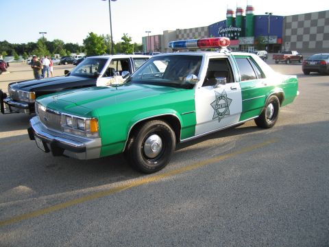 1991 Ford Crown Victoria - A green and white police car is parked in a lot next to another vehicle, with a movie theater in the background.