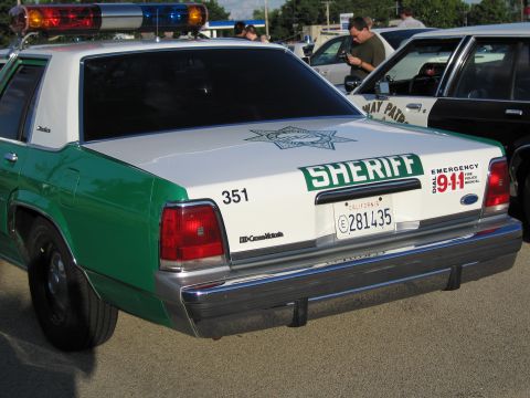 1991 Ford Crown Victoria - A Sheriff's patrol car is parked next to another vehicle, displaying emergency contact information on its rear.