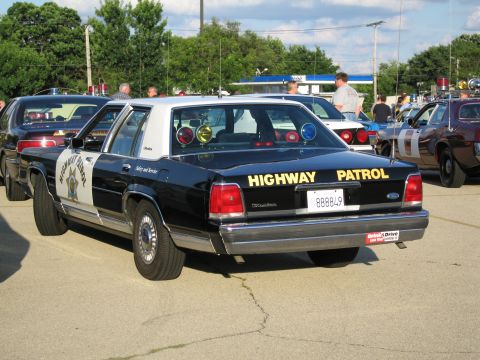 1991 Ford Crown Victoria - A black 1991 Ford Crown Victoria police car is parked outdoors with other vehicles and people in the background.