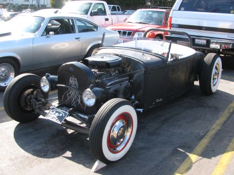 1990 Ford Bronco - flat-black open-wheel roadster hot rod with pinstriping and wide whitewall tires sits in a parking lot.