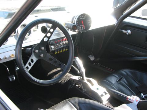 1990 Ford Bronco - stripped race-car interior shows a black three-spoke steering wheel, large tachometer, toggle switches and a black bucket seat.
