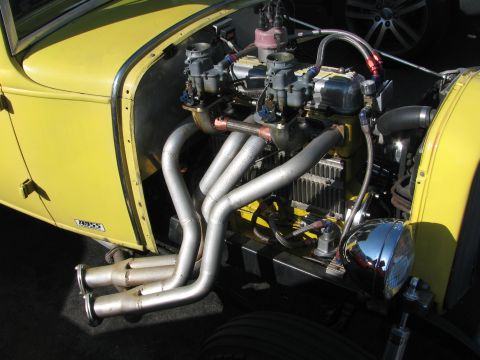 1990 Ford Bronco - close-up of a yellow hot rod's exposed V8 shows twin carburetors, finned valve covers and chrome zoomie headers.