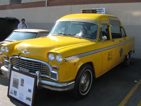 1990 Ford Bronco - yellow Checker-style taxi cab with a checkerboard stripe sits parked next to an informational display board.