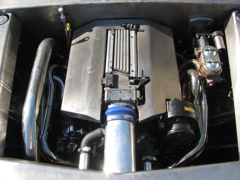 1990 Ford Bronco - clean engine bay shows a shrouded motor flanked by polished exhaust pipes and a blue intake hose inside a metal enclosure.