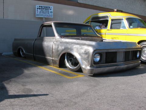1990 Ford Bronco - slammed classic pickup truck with bare unpainted bodywork and chrome wheels sits low to the ground beside a yellow vintage van.