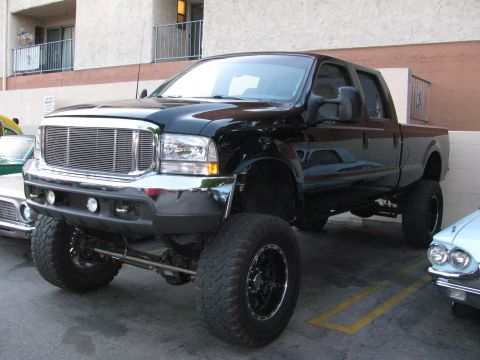 1990 Ford Bronco - lifted black Ford F-250 Super Duty crew cab pickup on black off-road wheels, parked in a lot beside other cars.