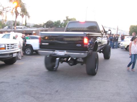 1990 Ford Bronco - heavily lifted black Ford Super Duty dually pickup driving through a crowded parking lot at dusk among onlookers.