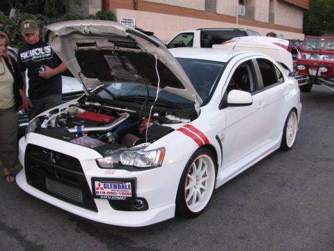 1990 Ford Bronco - white Mitsubishi Lancer Evolution with red striping and open hood at an outdoor car show, with people looking on.